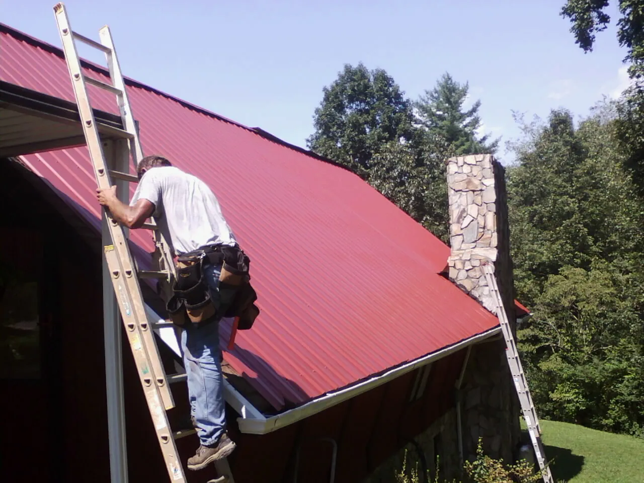 Roofer on a ladder beside a red metal roof and stone chimney — Soffit & Fascia Repair by Roofing Brothers in Meridale